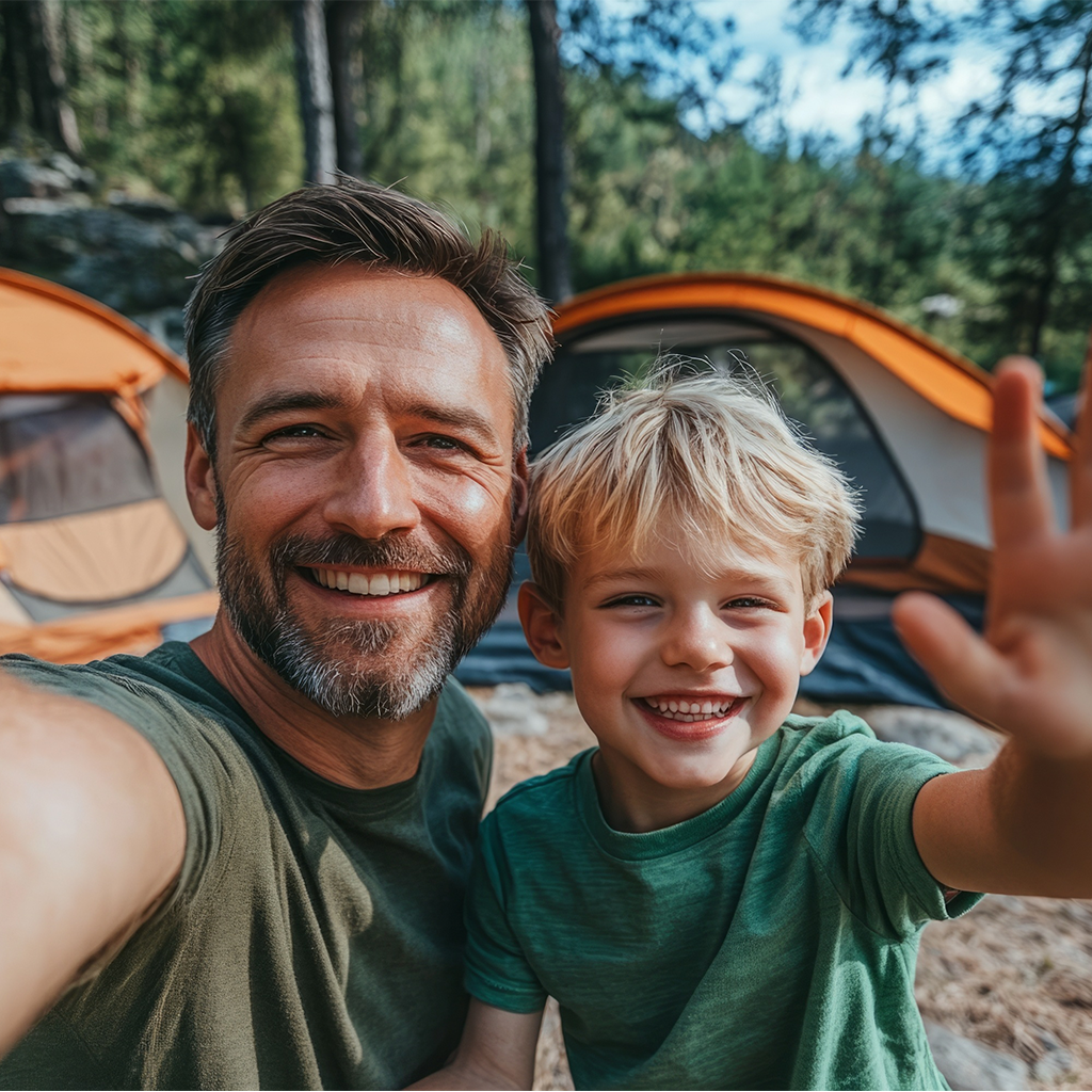 Täiskasvanu ja laps telkimisel selfit tegemas, taustal mets ja telgid